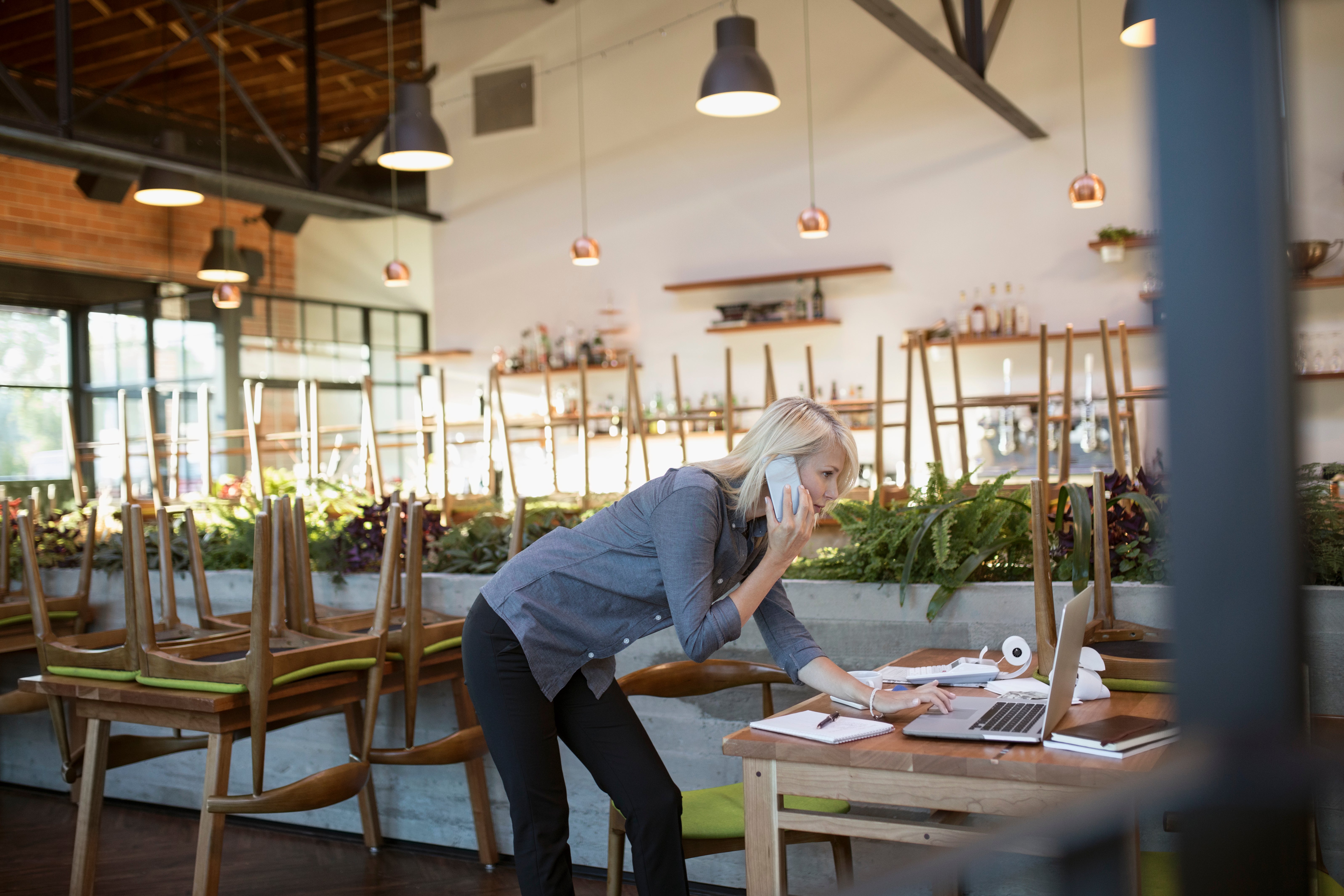 Female cafe business owner talking on cell phone, working at laptop