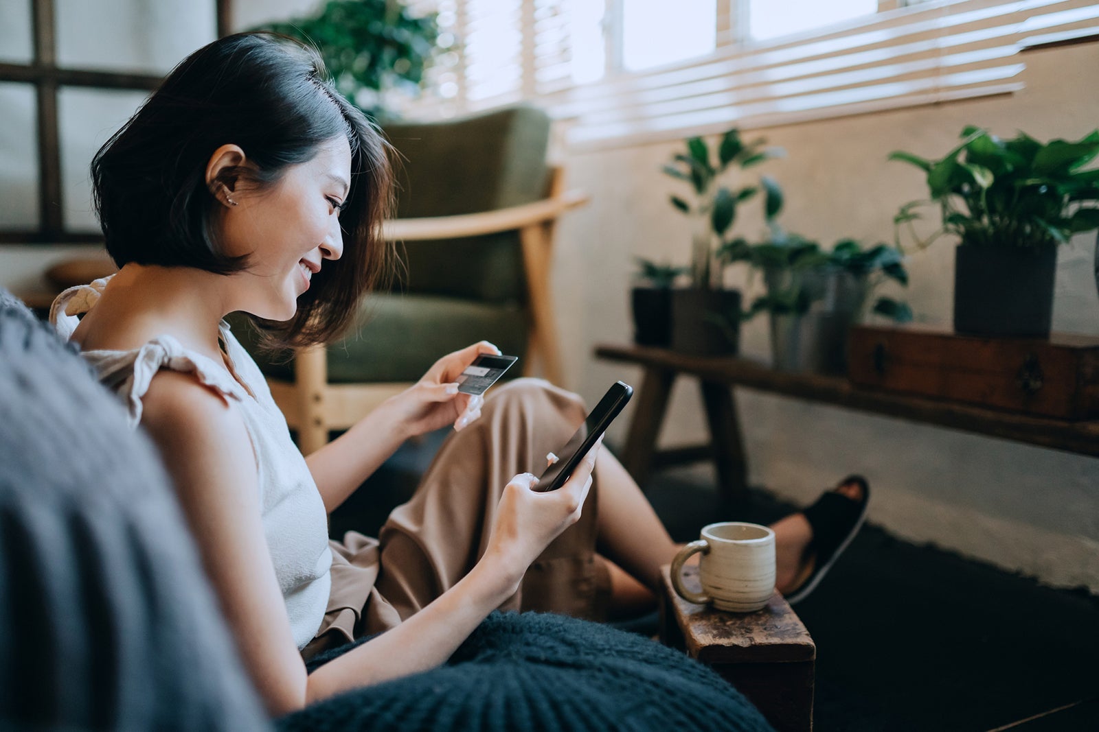 Beautiful smiling young Asian woman chilling at home, sitting on the floor in bedroom, enjoying a cup of coffee and shopping online with smartphone while making mobile payment with credit card on hand