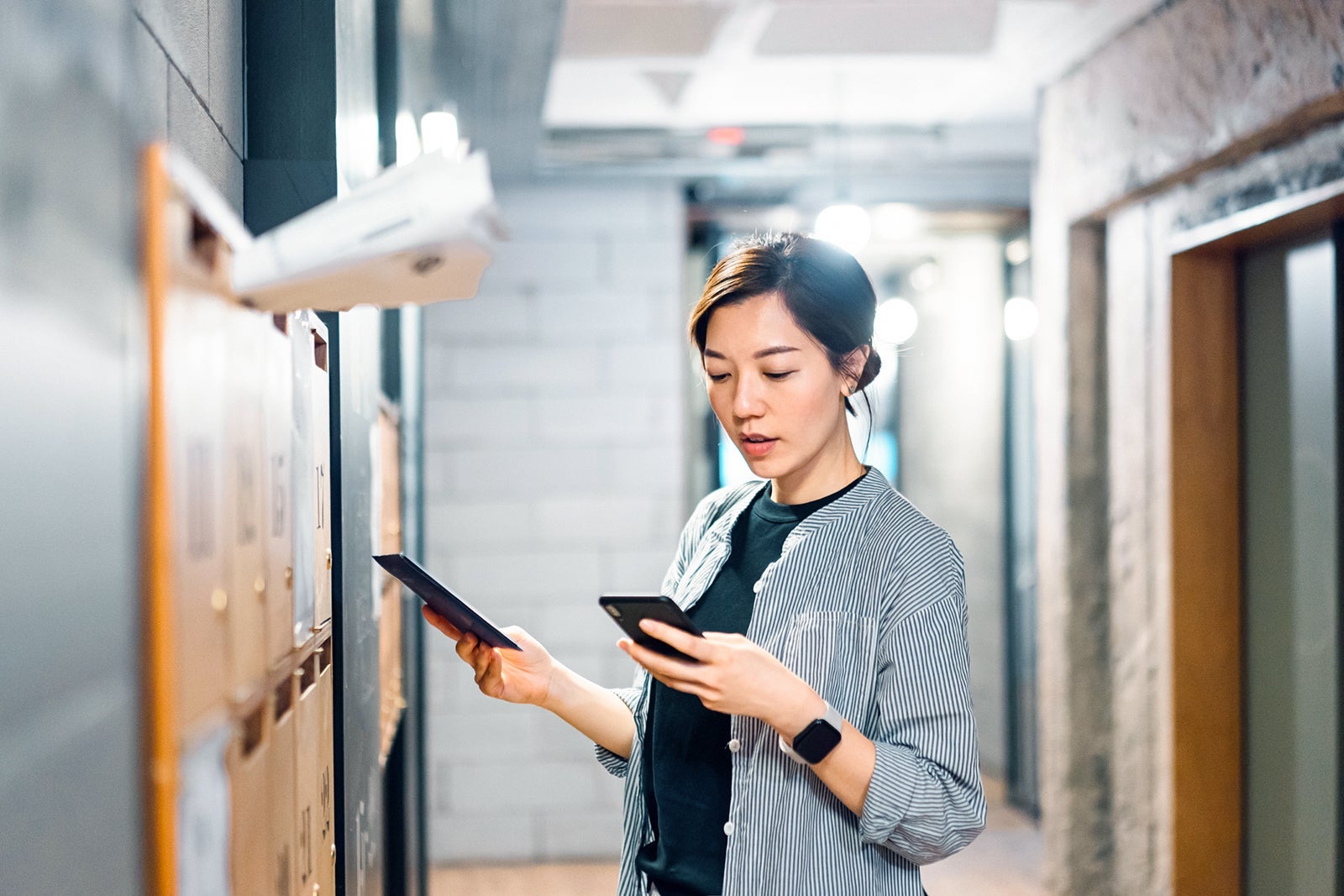 Young Asian female start-up business owner checking smartphone while collecting mails from mailbox in her shared office