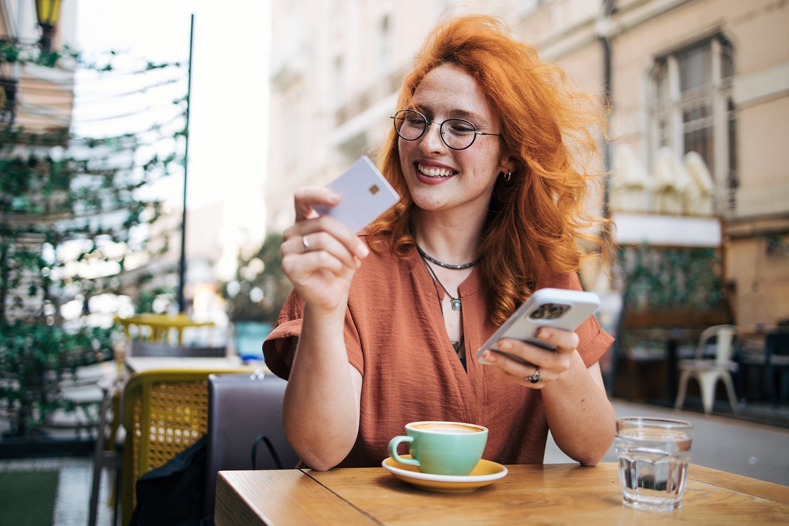 Happy young redhead woman in cafe drinking coffee and shopping online on smart phone